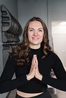 Woman with hands in prayer pose smiles. She wears black clothing against a gray and white background.