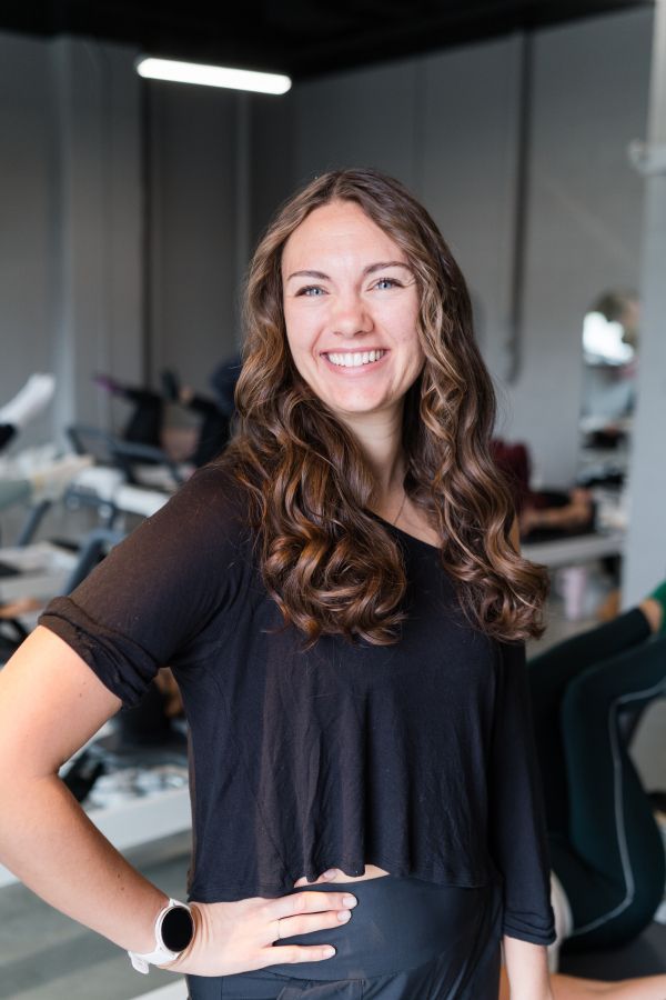 Woman with hands in prayer pose smiles. She wears black clothing against a gray and white background.