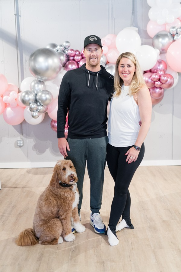 A man and a woman are standing next to a dog in front of balloons.