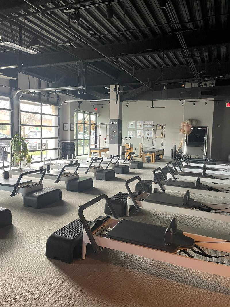 Pilates studio with rows of reformer machines. Gray flooring, dark ceiling, and large windows.