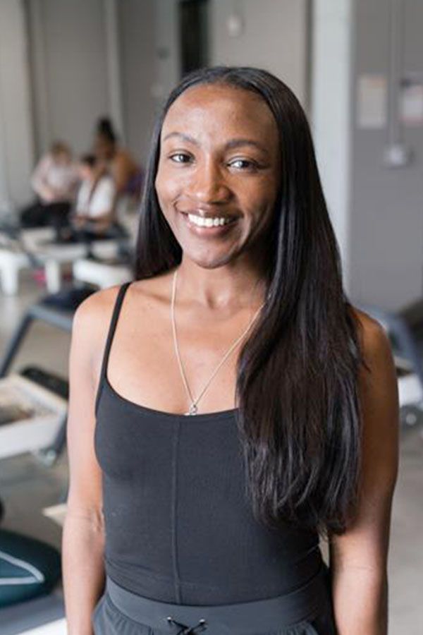 Woman smiling in a black tank top and leggings, standing in a gym with exercise equipment