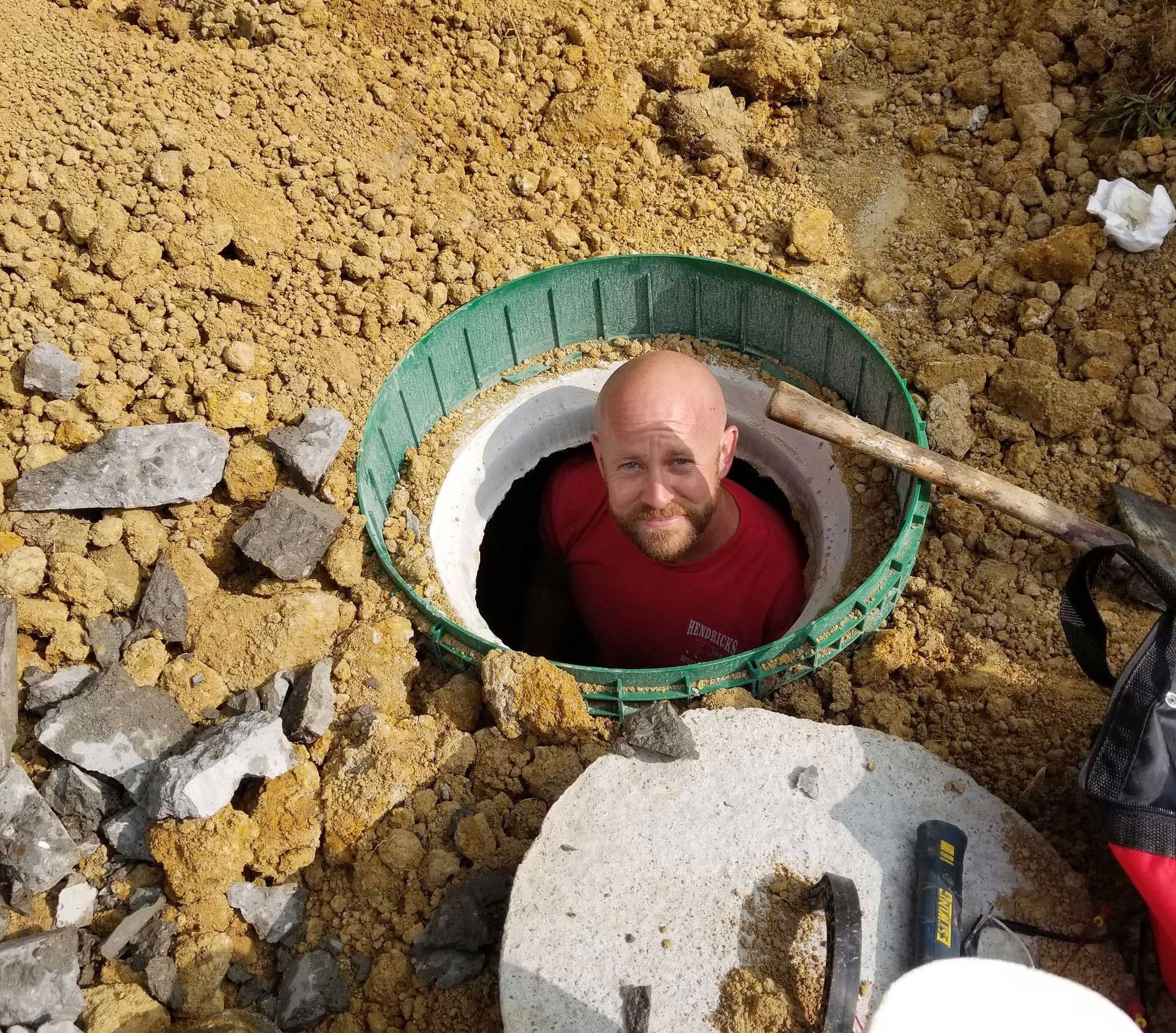 Man in red shirt inside a septic tank, surrounded by dirt and debris.