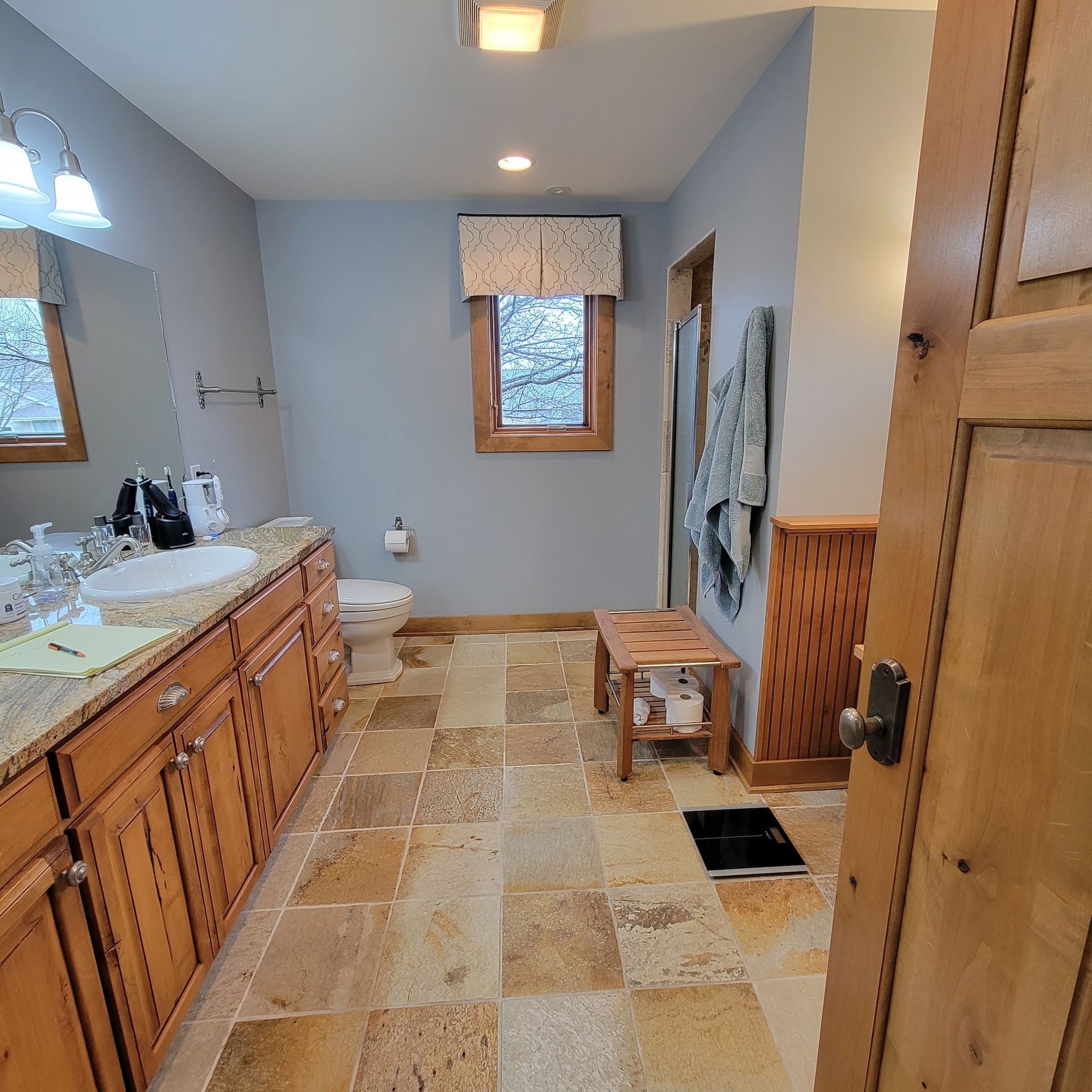 Bathroom with blue walls, wooden cabinets, stone floor, and a window with a curtain.