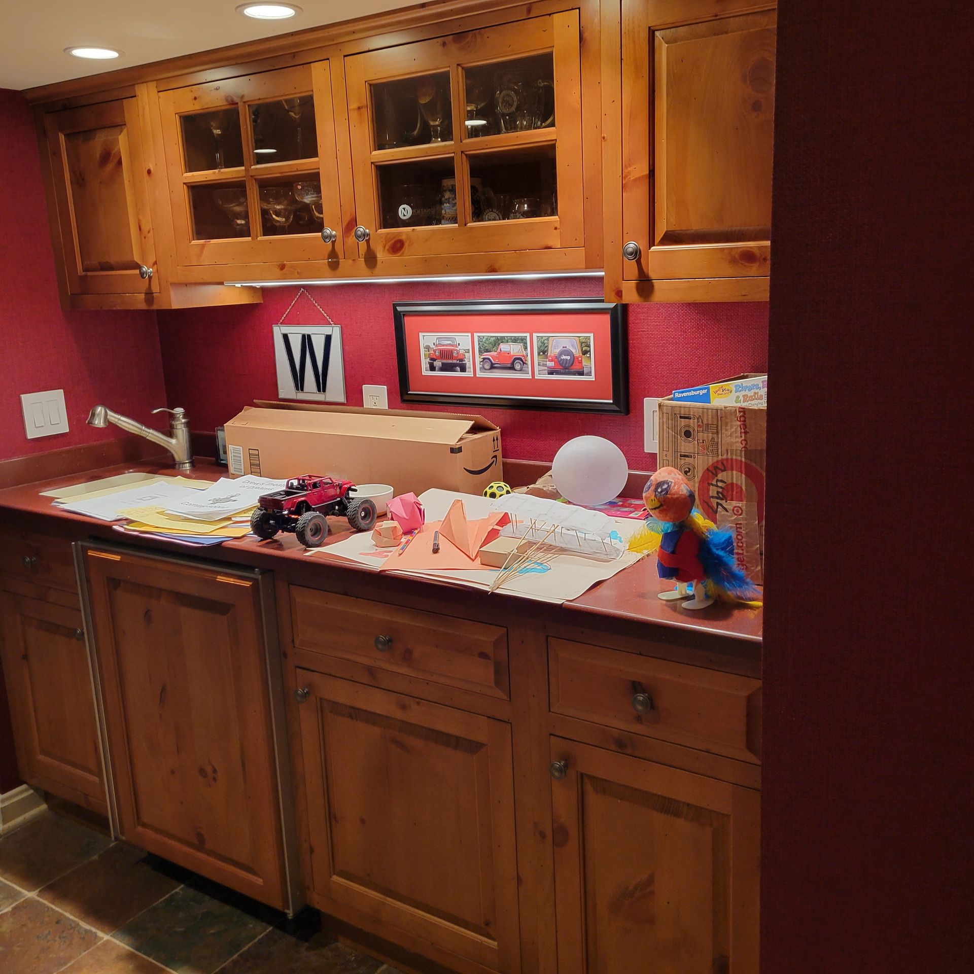 Wooden cabinets and countertop with craft supplies and toys against a red wall.