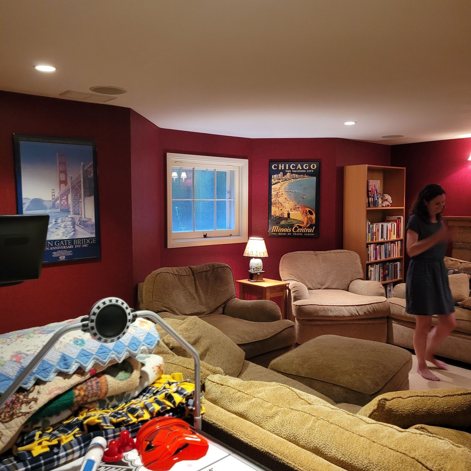 Woman in a living room with red walls, beige couches, and a bookcase. A baby play gym is in the foreground.