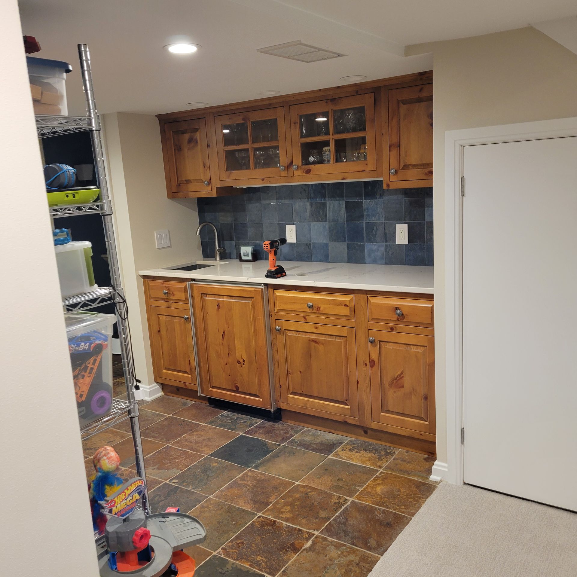 A basement bar with wooden cabinets, tile backsplash, and stone flooring.