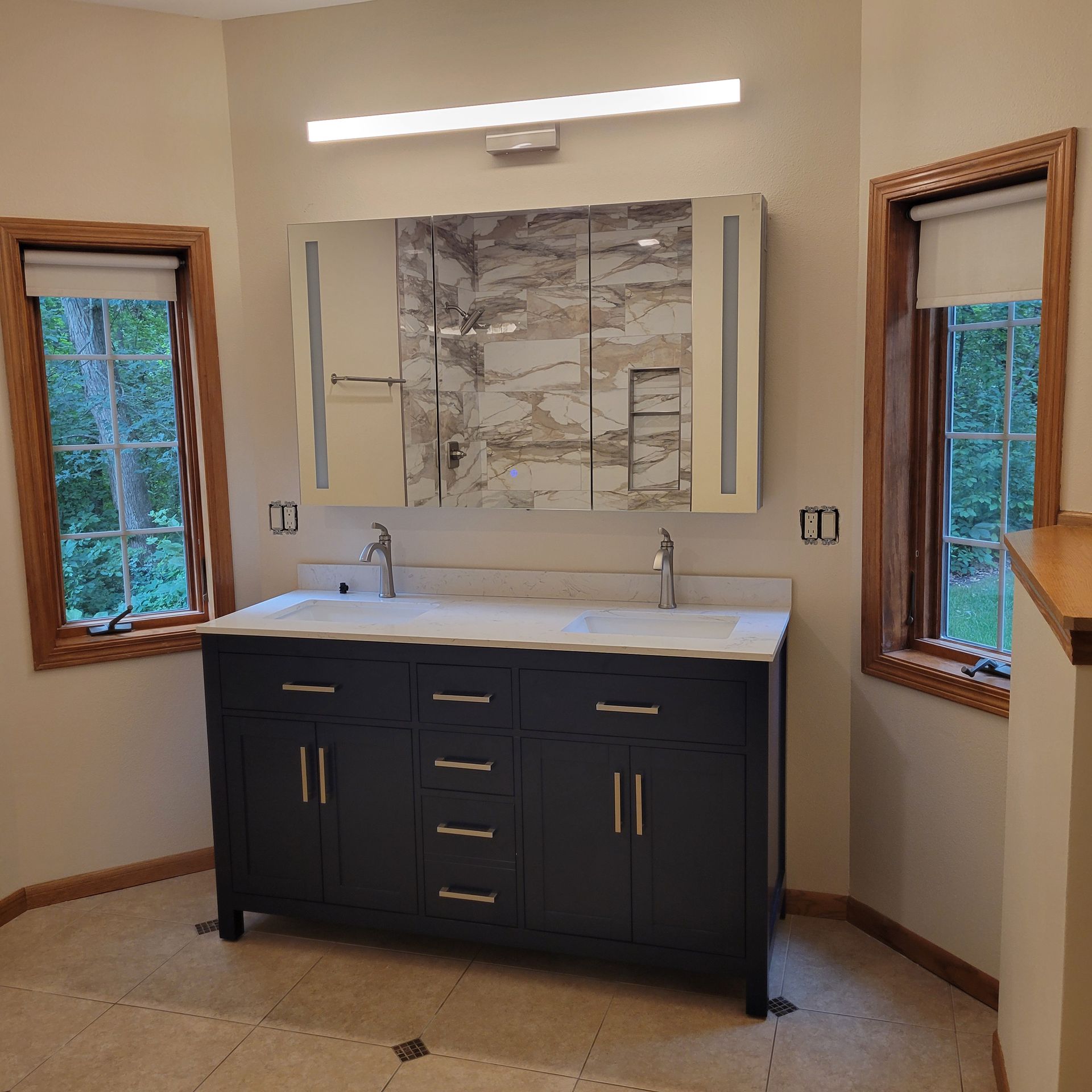 Bathroom with dark blue vanity, double sinks, large mirror with lights, and two windows.