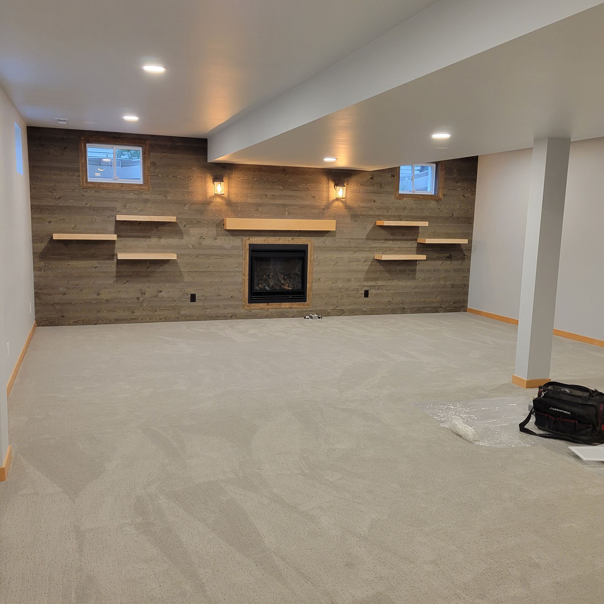 Finished basement with fireplace, shelves, and neutral carpet. Light fixtures add warmth to the space.