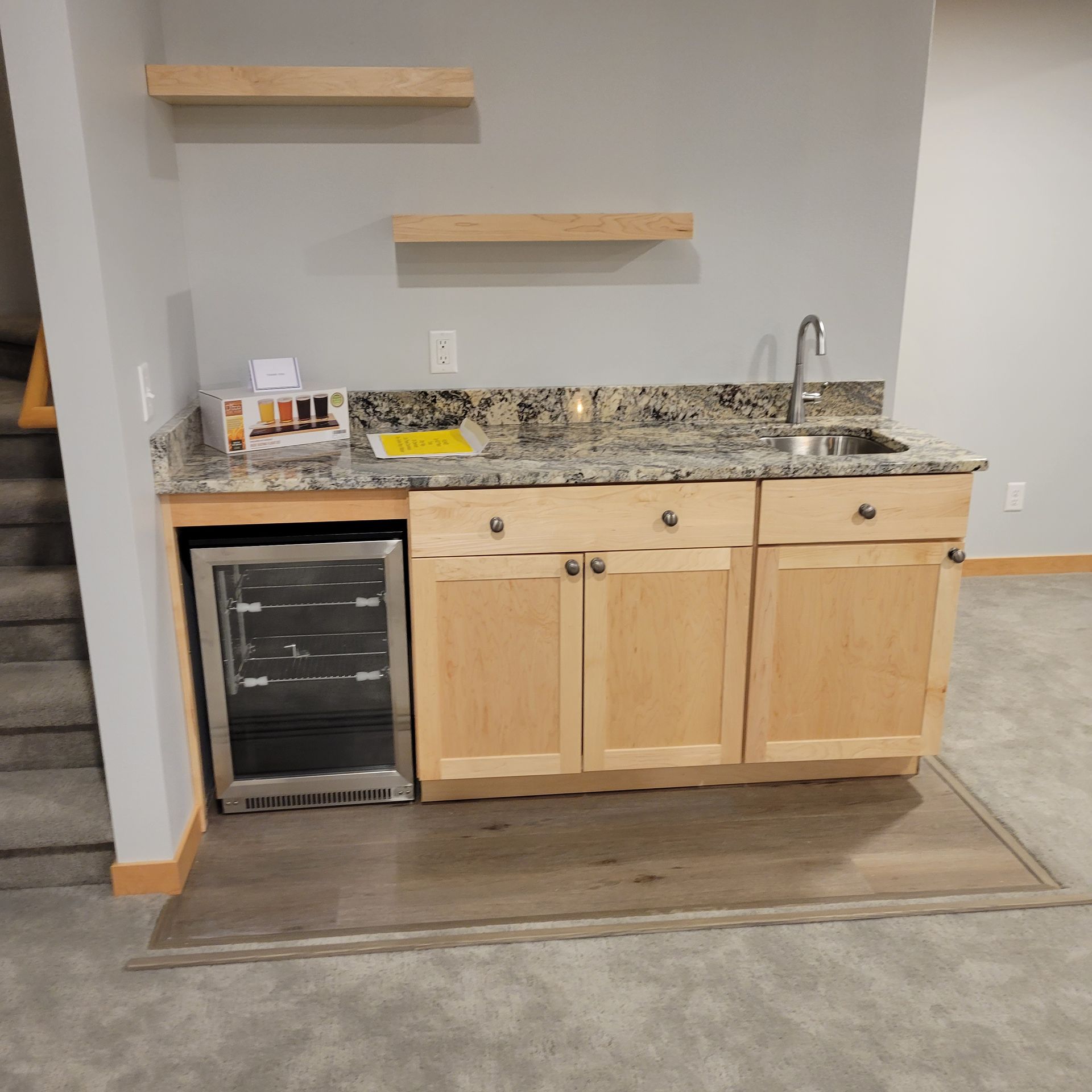 Basement wet bar with a small fridge, cabinets, granite countertop, sink, and shelves.