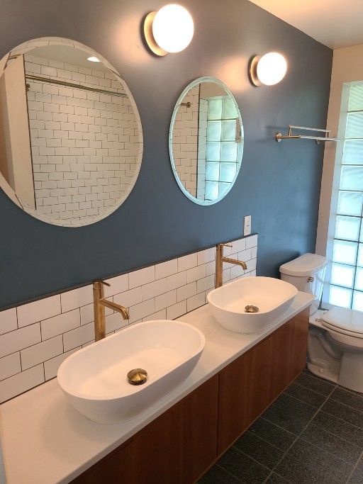 Modern bathroom with dual vessel sinks, round mirrors, blue walls, and gold fixtures.