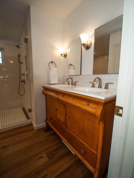 Bathroom with wooden vanity, marble countertop, double sinks, shower, hardwood floor, and arched mirror.