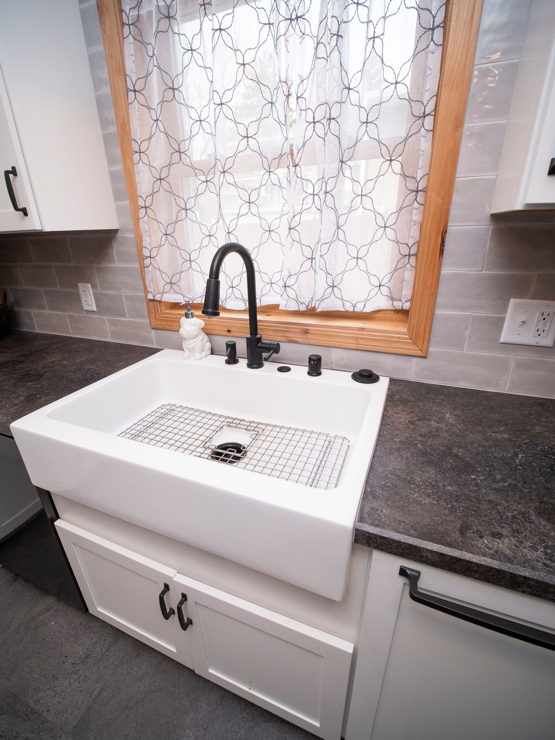 White farmhouse sink with black faucet, under a window with a patterned curtain.