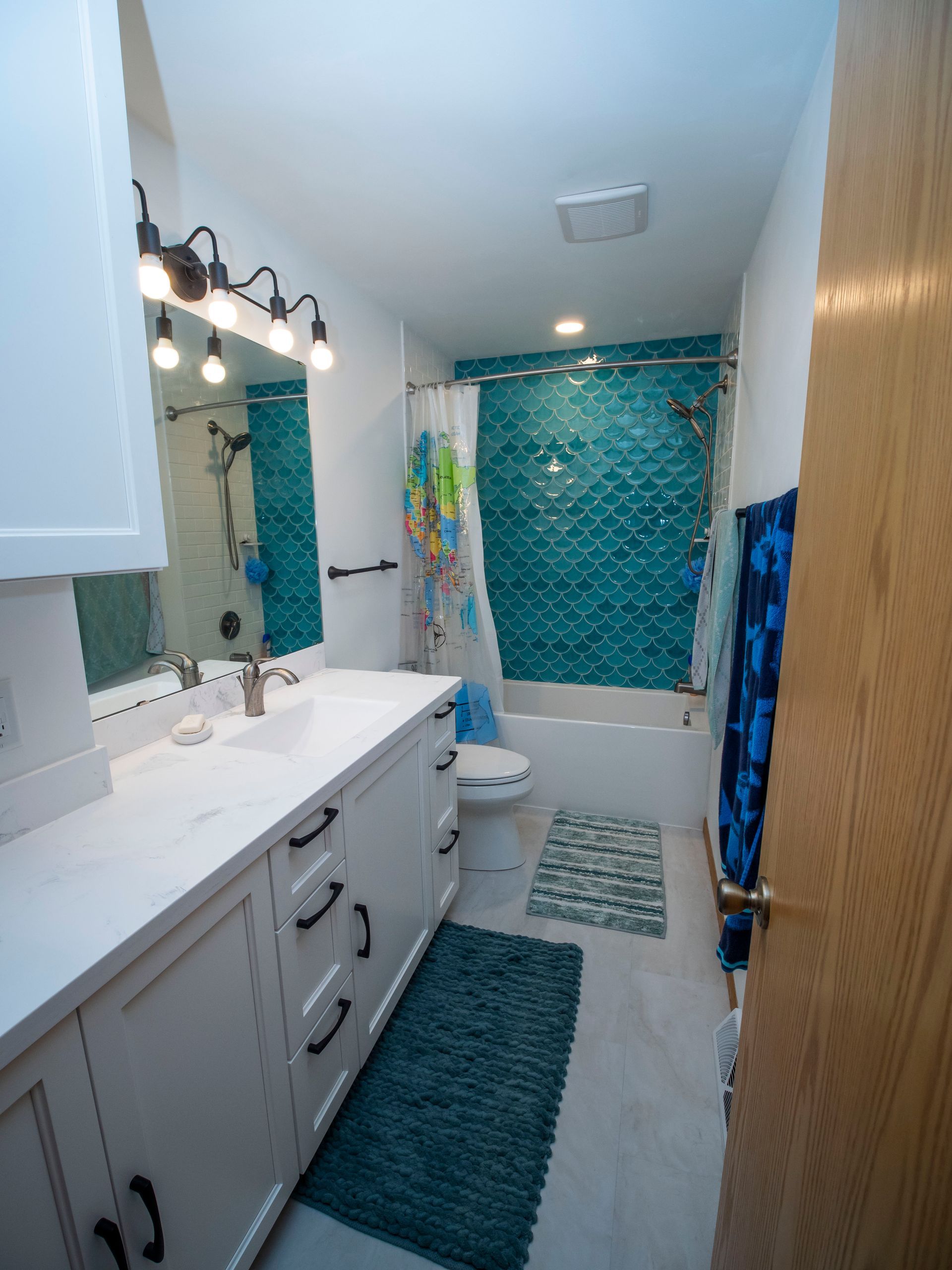 White bathroom with teal tile accent wall, long vanity, and light blue rugs.