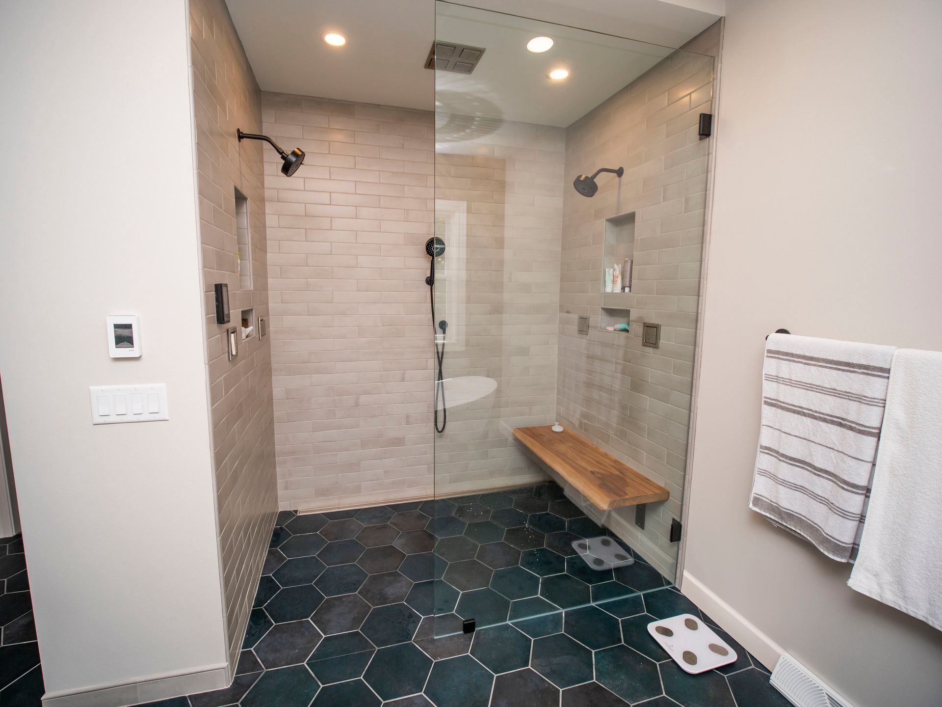 Spacious bathroom with walk-in shower, hexagon tile floor, wood bench, two shower heads, and glass door.