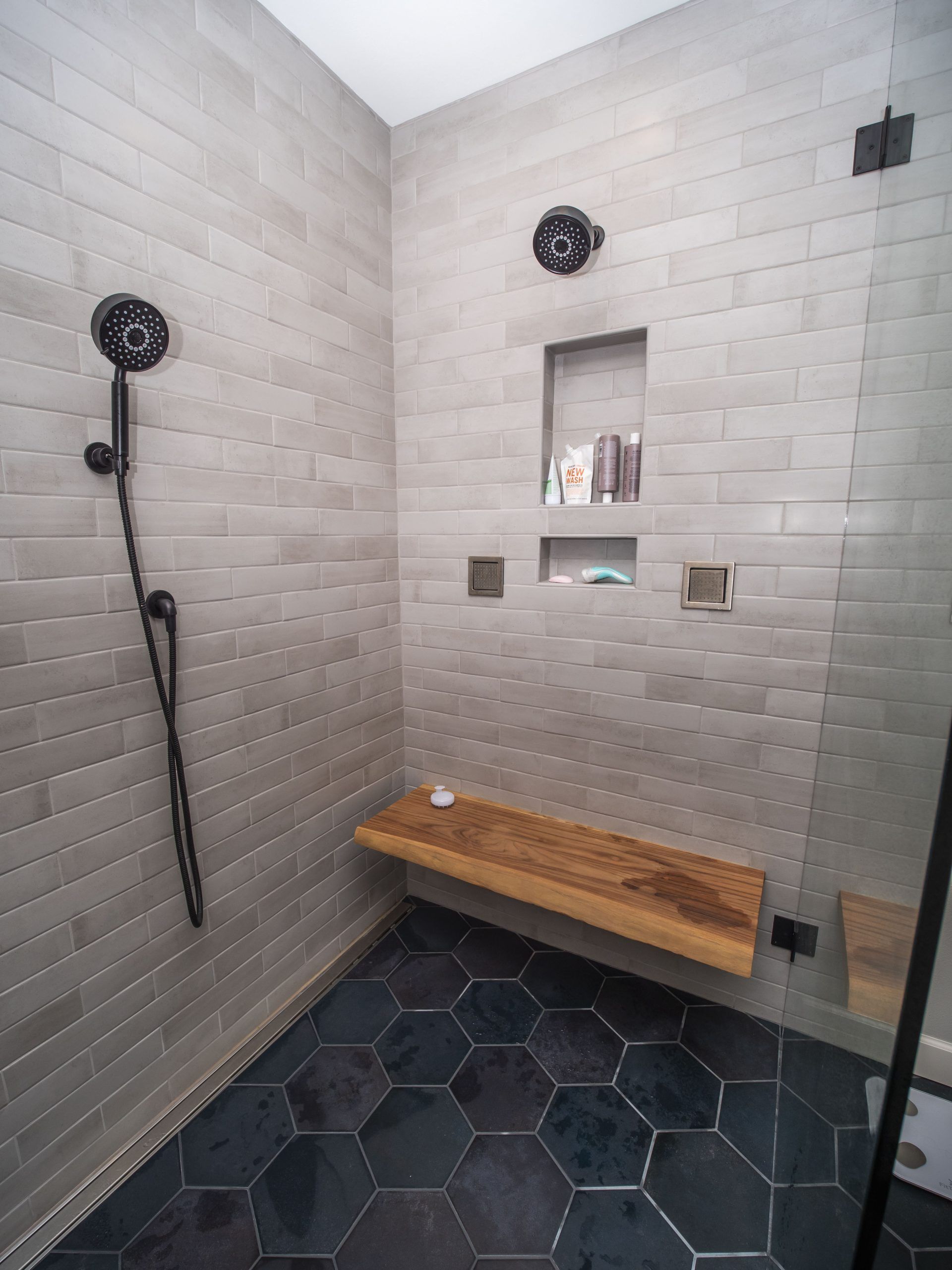 Modern shower with gray tiled walls, dark hexagon floor, wooden bench, black fixtures.