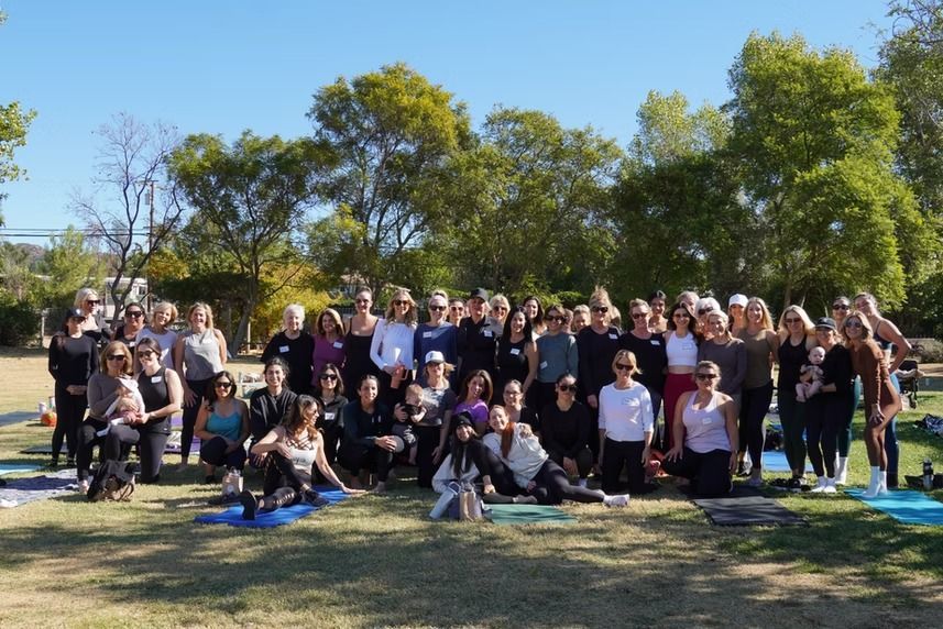 Group of people posing outdoors on a grassy field with trees and a bright blue sky.