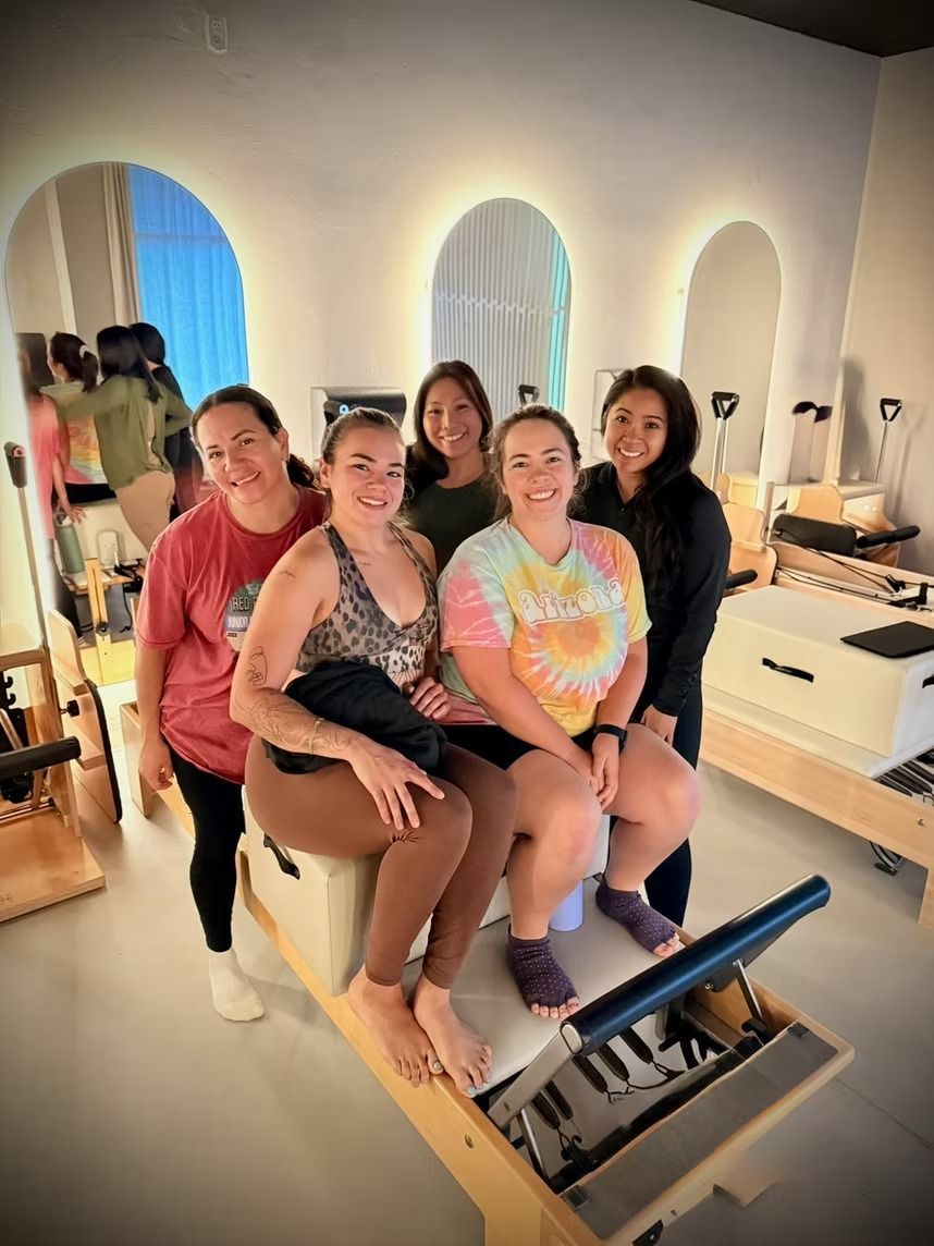 Four people smiling on a Pilates chair in a bright studio, with mirrors and arched windows behind them.