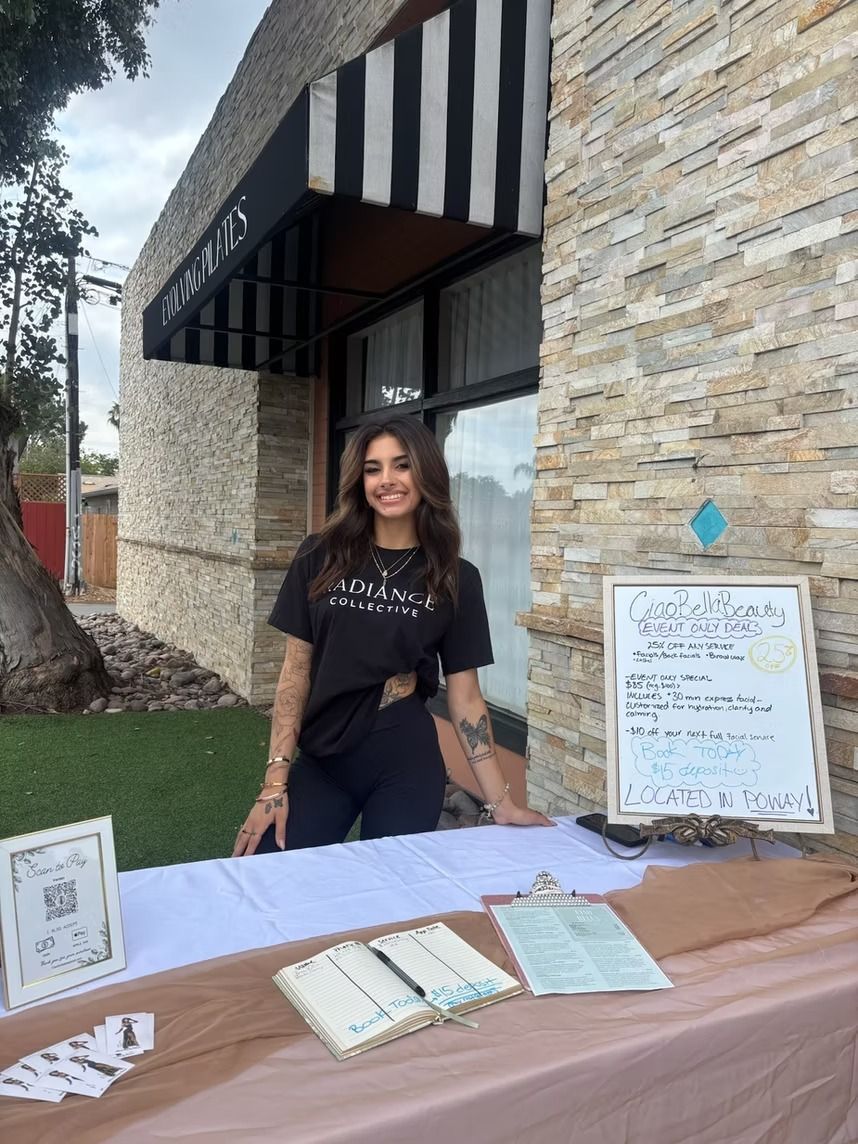 Woman standing behind an outdoor vendor table beside a stone building with a striped awning and signboard.
