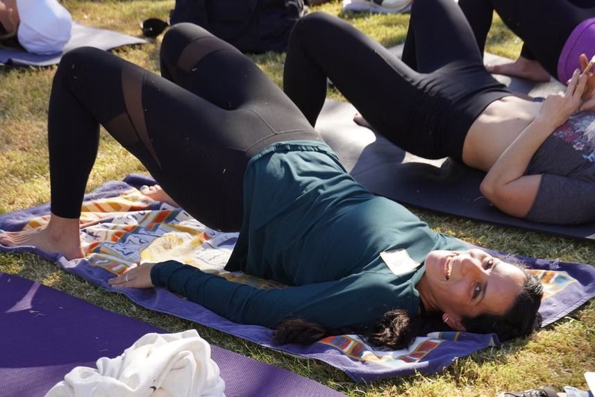 People lying on mats outdoors, doing a bridge yoga pose in a sunny park