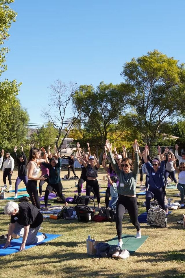 People exercising outdoors on yoga mats in a park, arms raised under a clear blue sky.