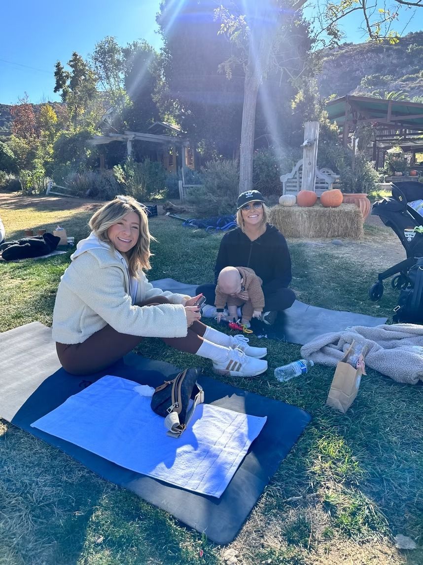 Two people sitting on blue mats outdoors in sunlight with a small black dog between them.