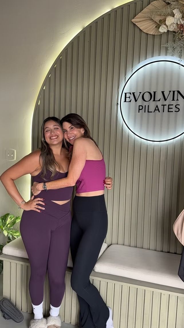 Two women posing together in matching workout outfits in front of an Evolvin Pilates sign.