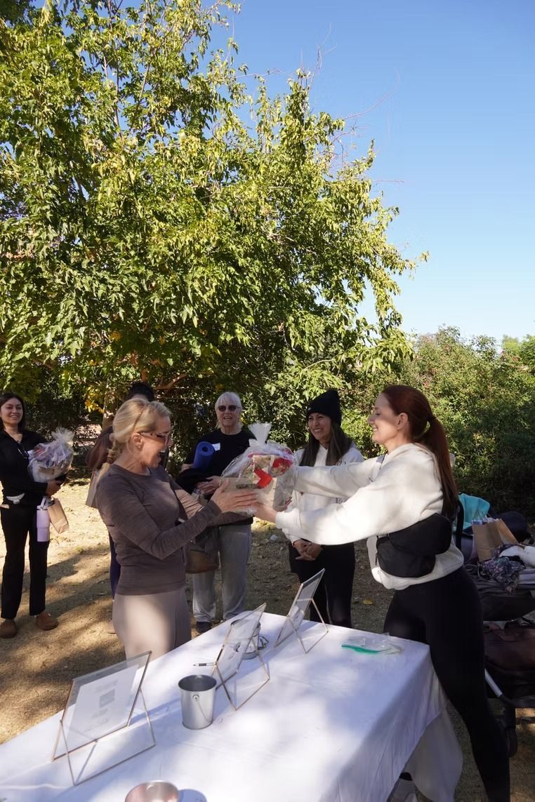 People exchanging a wrapped gift outdoors beside a table under a tree, with others watching.