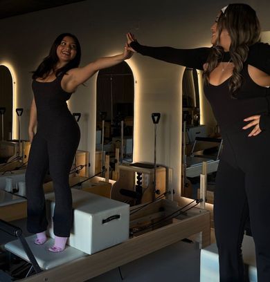 Two women in black outfits pose on Pilates reformers, touching hands in a studio.