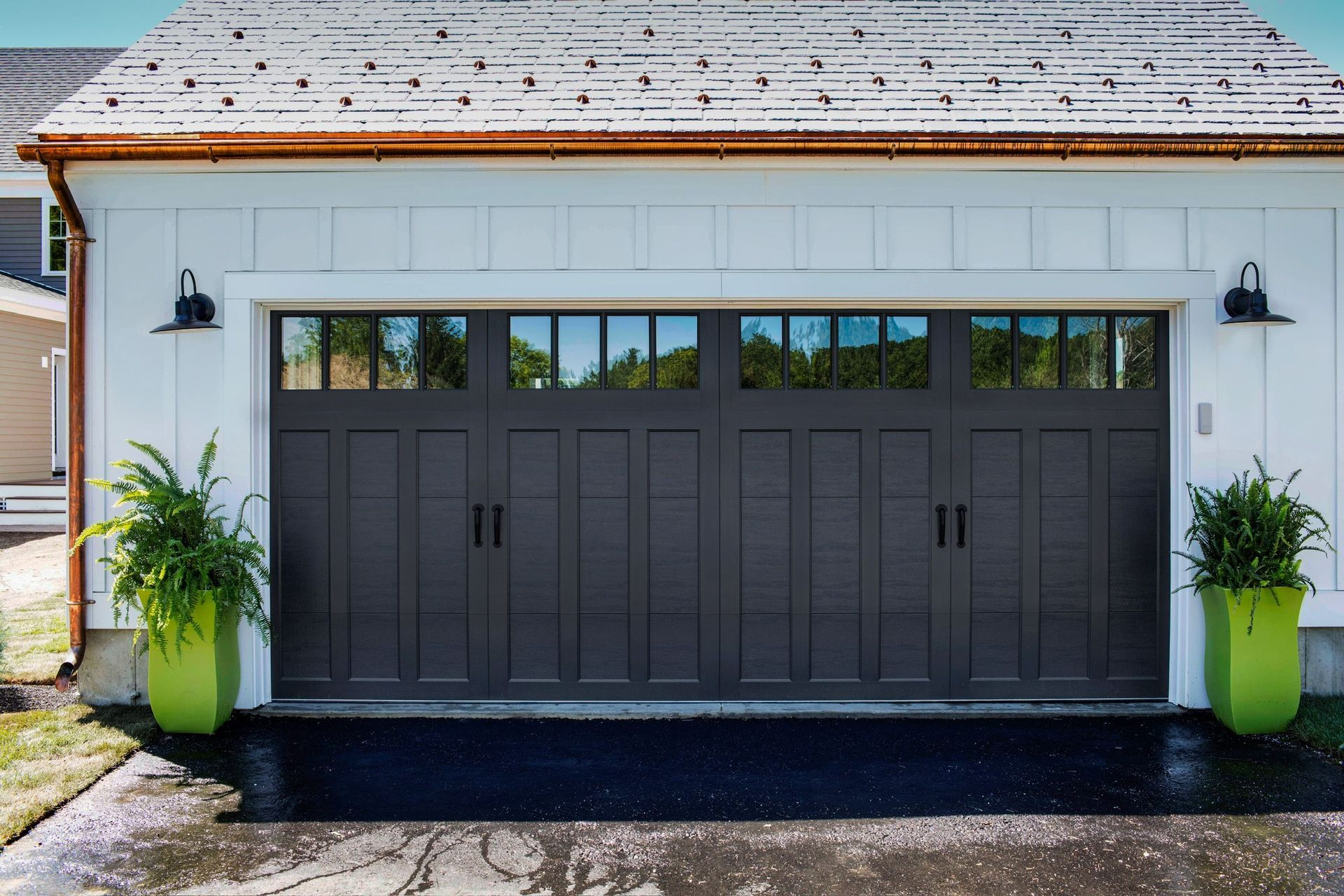 A white house with a black garage door and green potted plants.