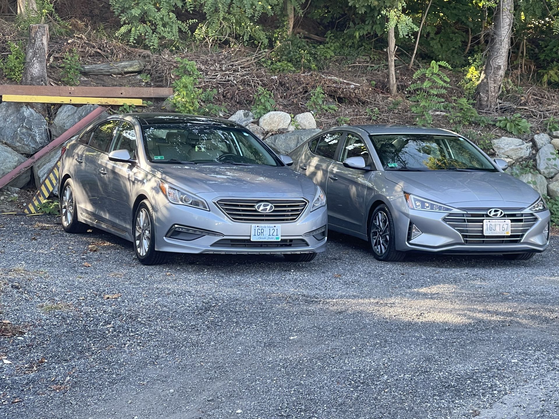Two gray cars are parked next to each other in a gravel lot.