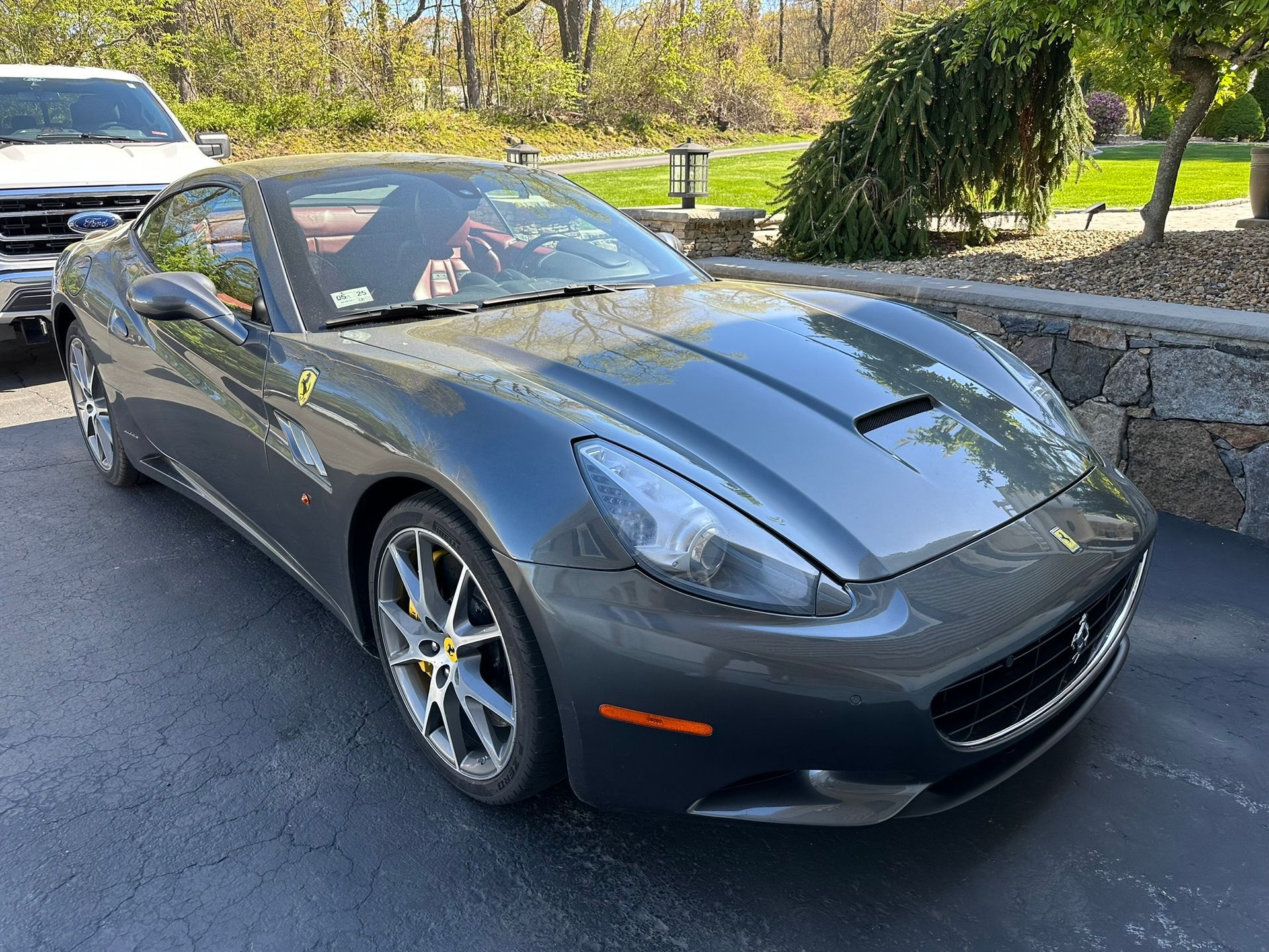 A gray ferrari is parked in a driveway next to a white truck.