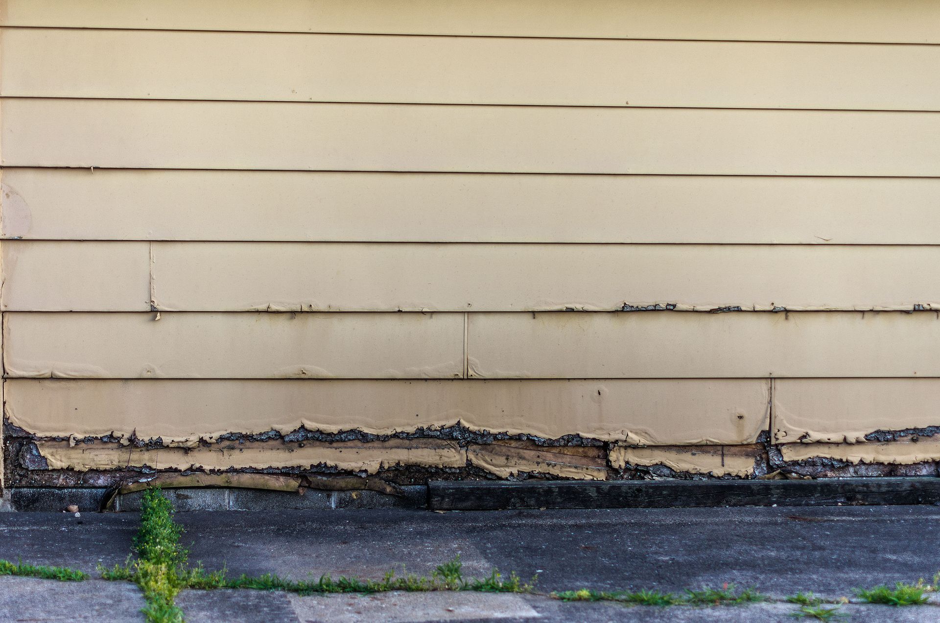 Weathered beige house siding with peeling paint above a dark, cracked foundation and grass at the base