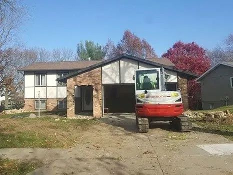 Excavator parked in front of a house with an open garage, framed by autumn trees.