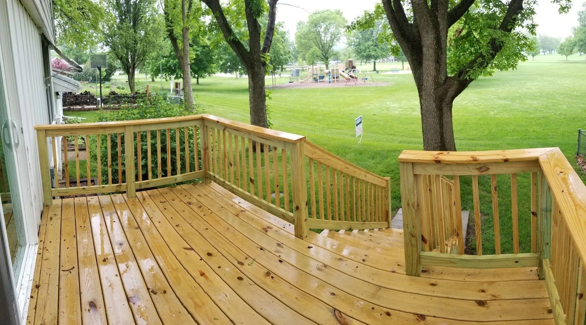 Wooden deck with railing overlooking a green park with trees and open grass.