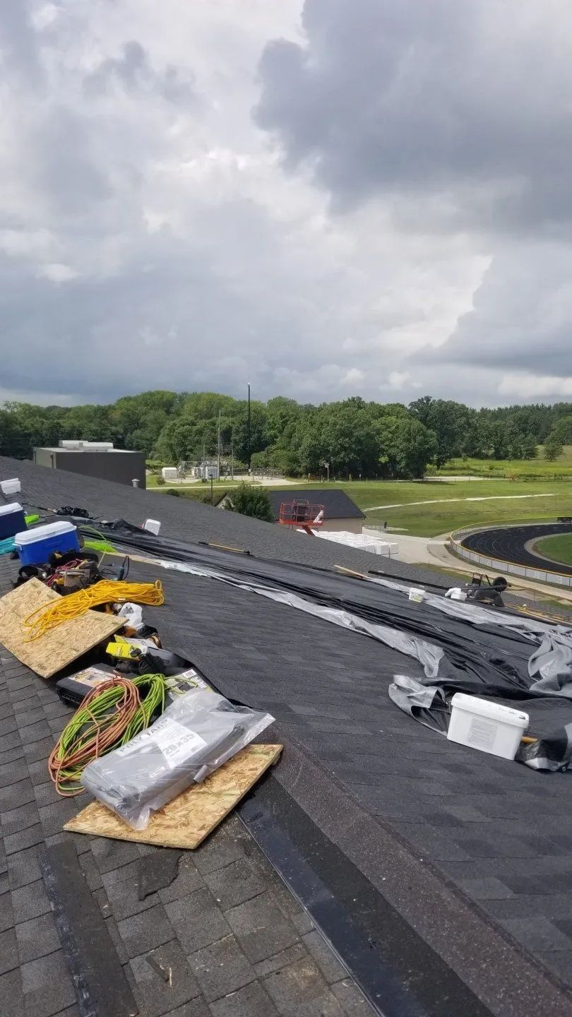 Rooftop under repair with workers’ materials, gray shingles, and cloudy sky over a green landscape