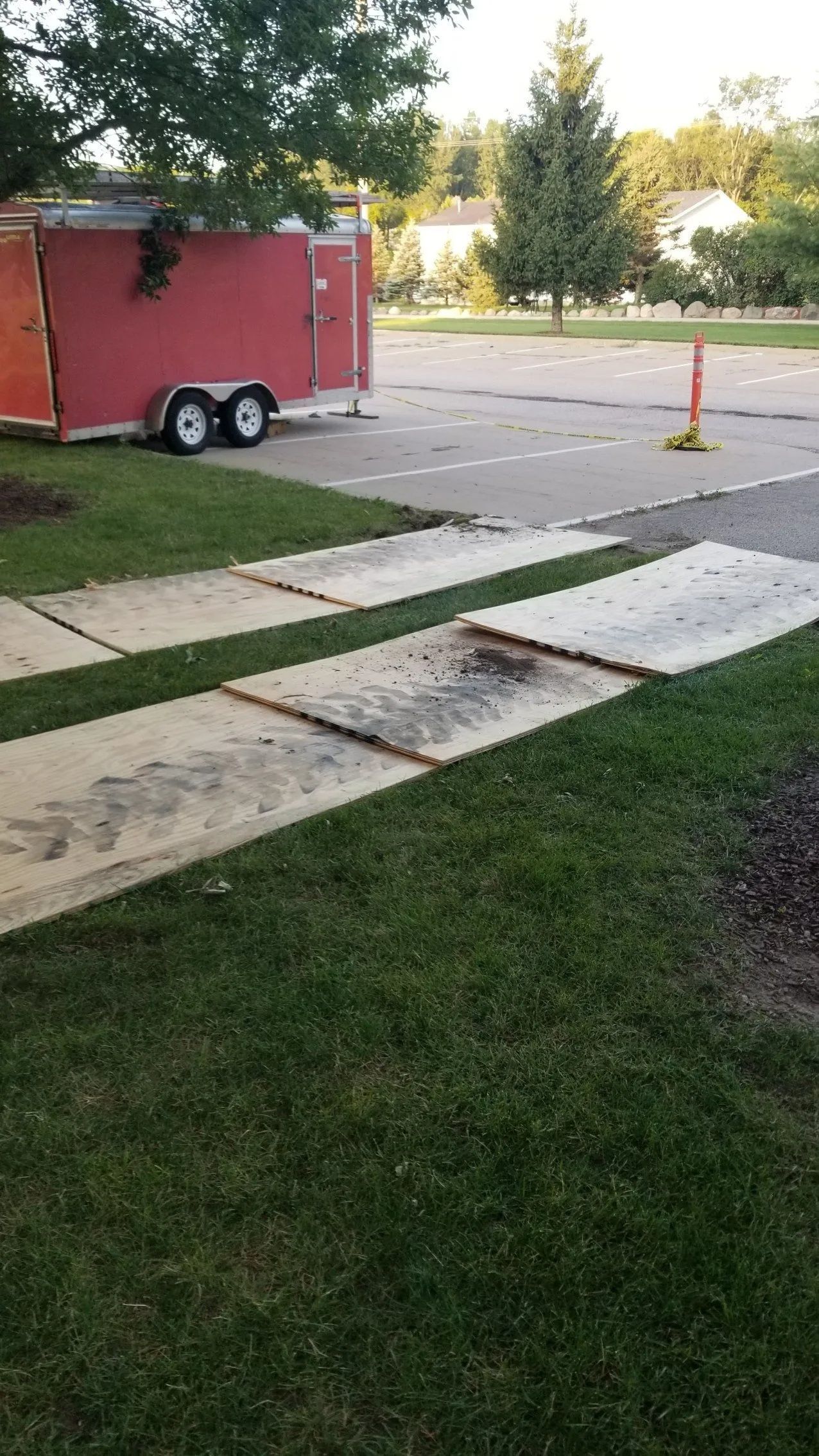 Broken sidewalk slabs beside a red trailer, with a grassy lawn and cones in a parking area.