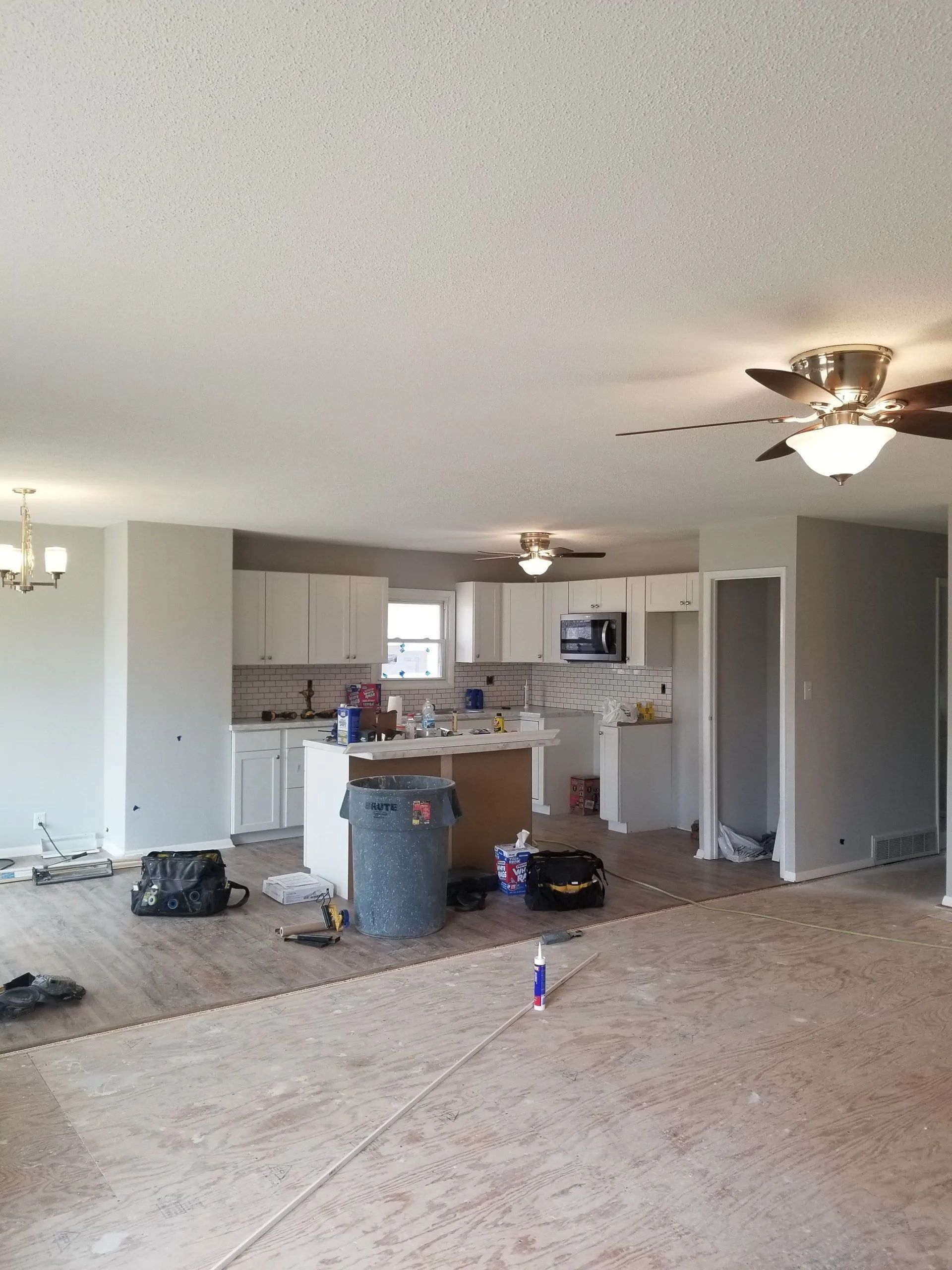 Unfurnished living room with exposed flooring, ceiling fans, and a cluttered kitchen in the background.