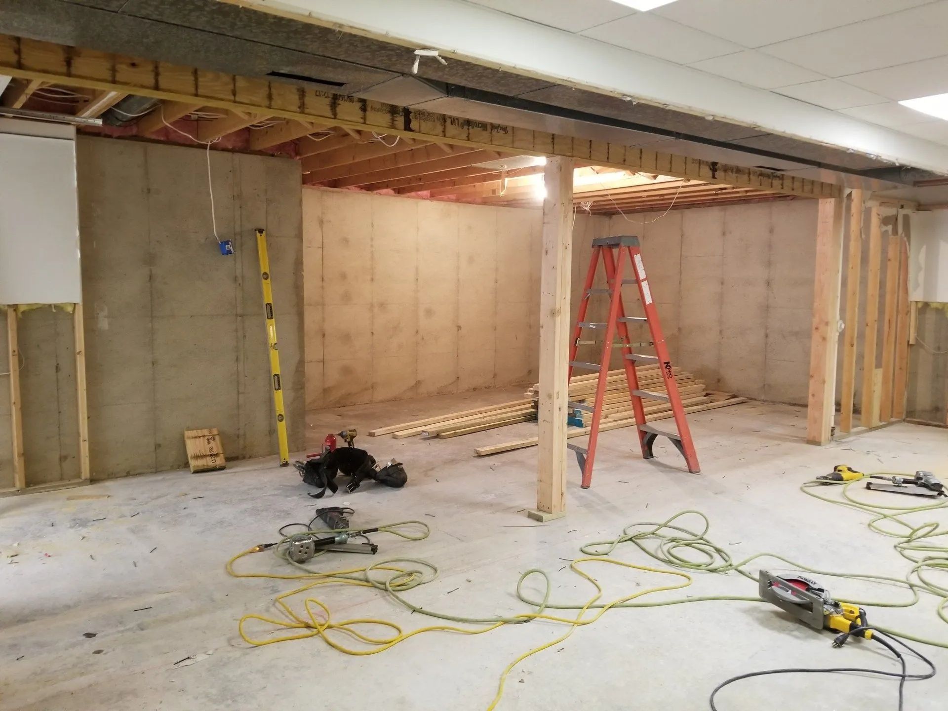 Renovation site with exposed wall framing, ladder, tools, and extension cords on a concrete floor