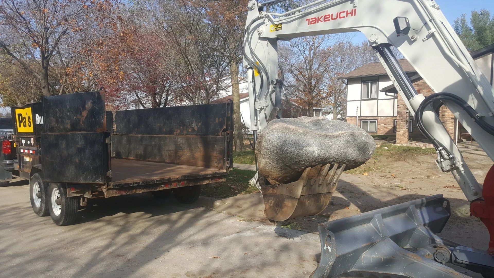 Excavator loading a large stone into a dump trailer on a roadside in autumn