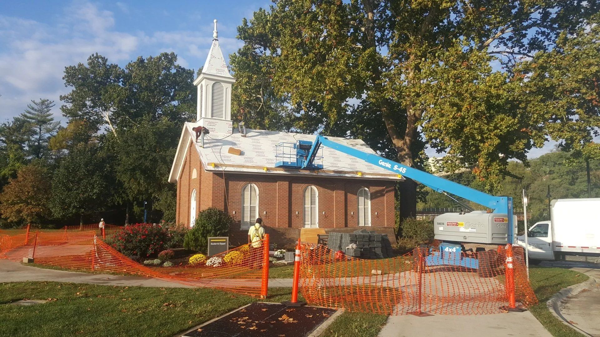 Small brick church under repair with blue roof tarps, orange fencing, and a bucket lift in front.
