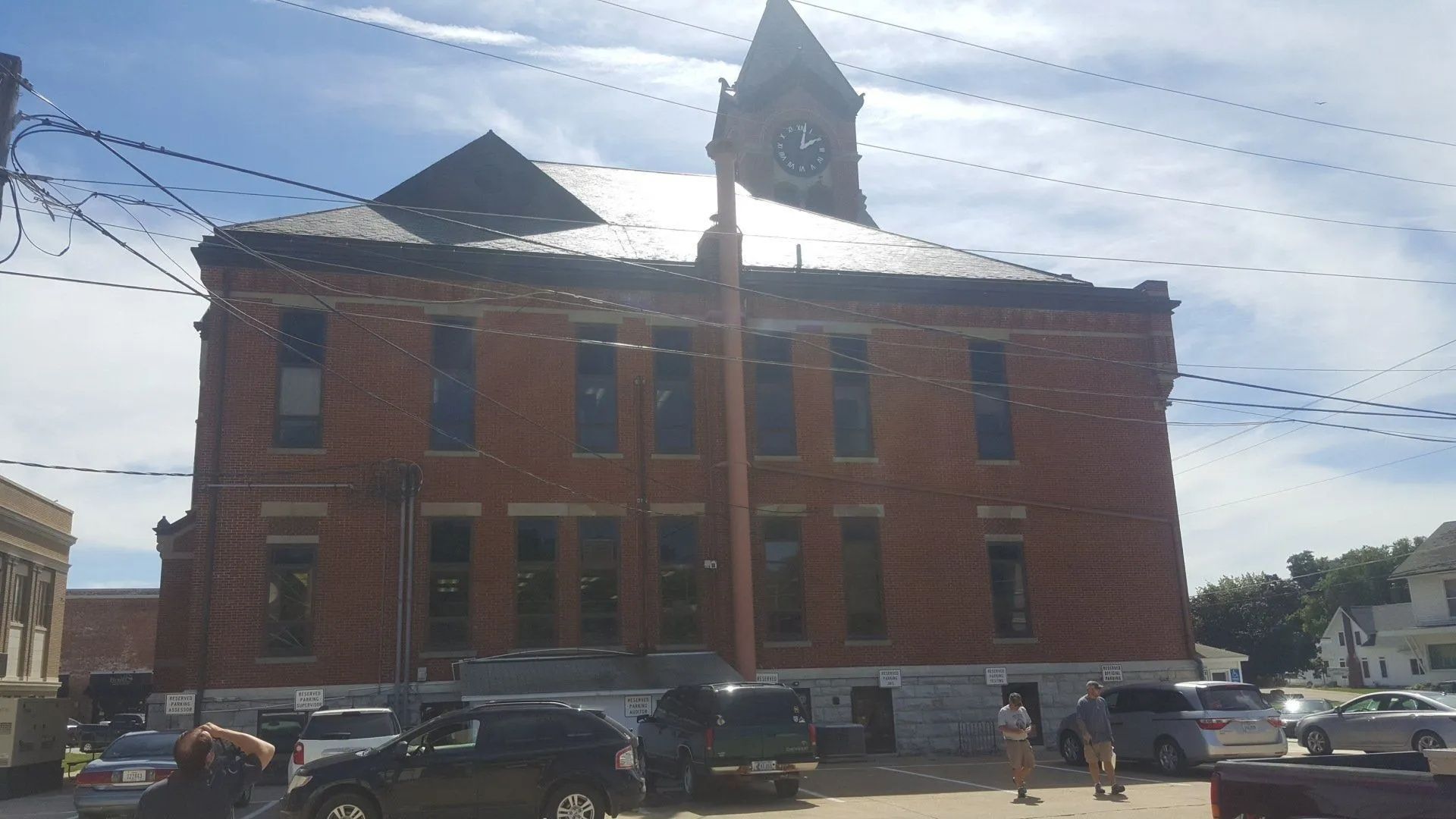 Brick building with a small tower, parked cars, and power lines on a sunny street