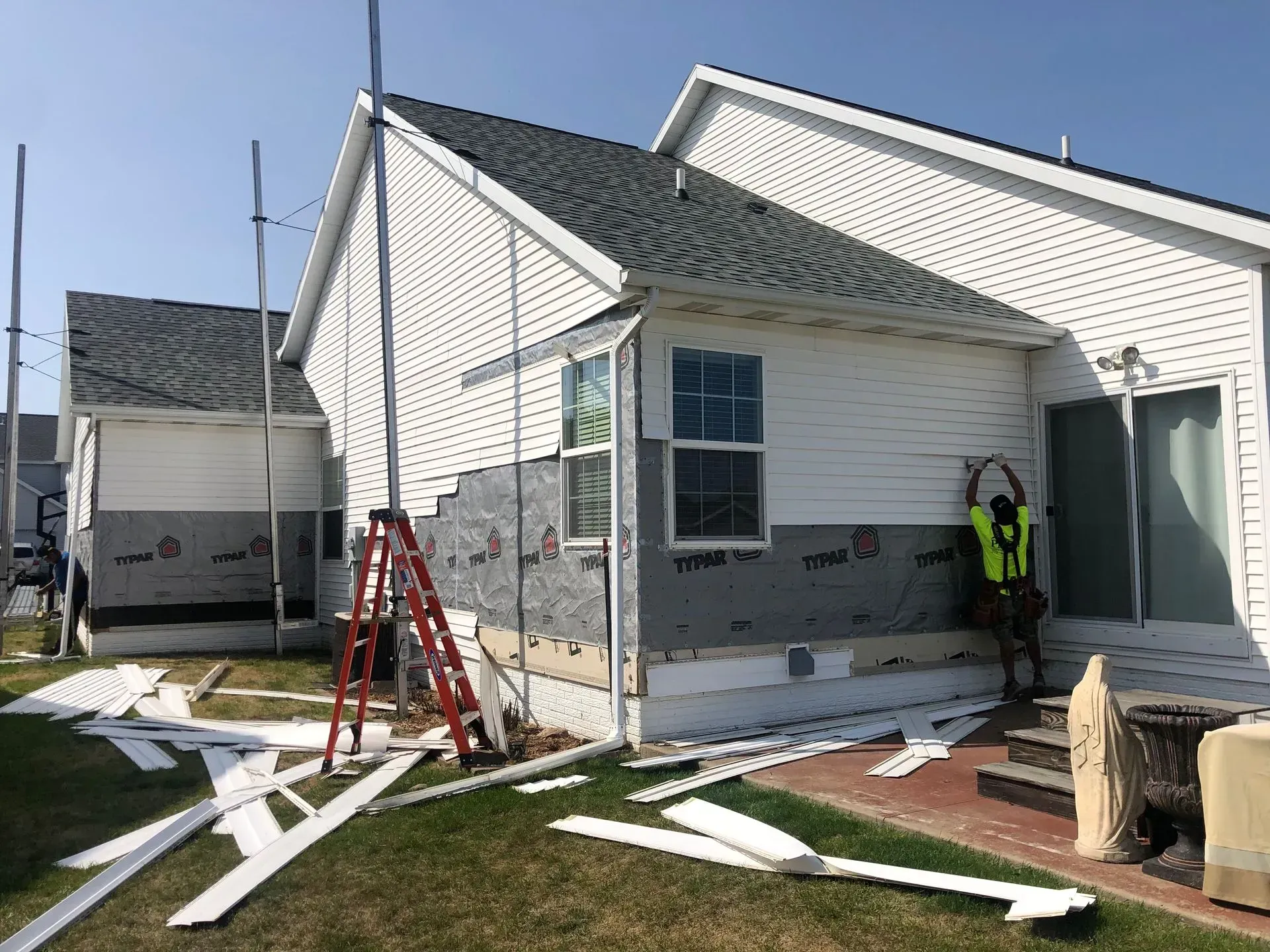 Workers in neon vests at a house under construction, with ladders, siding, and debris on the ground.