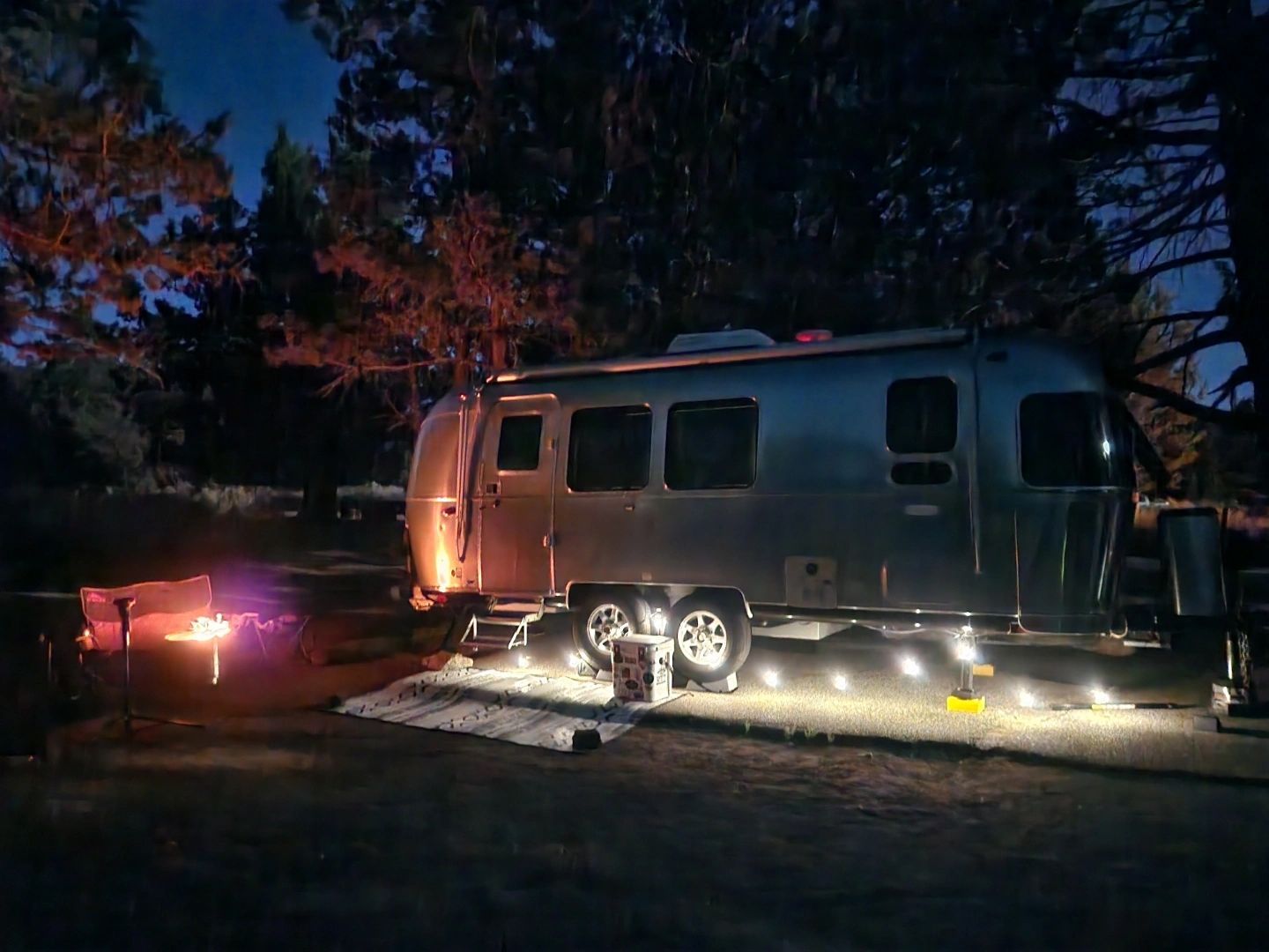 Silver Airstream trailer at night, illuminated by string lights. A campfire glows nearby.