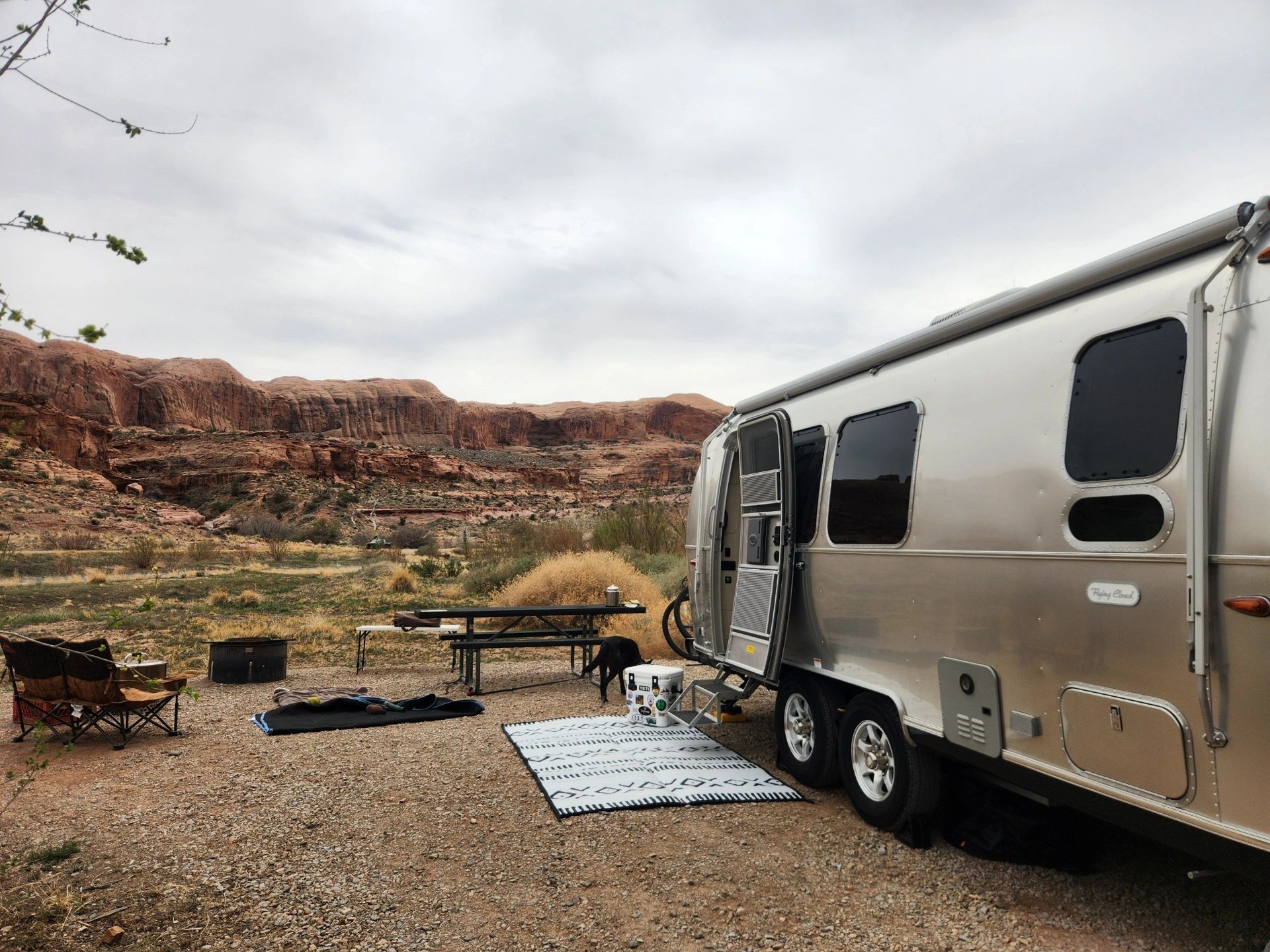 Airstream trailer parked in a campsite, overlooking red rock formations under a cloudy sky.
