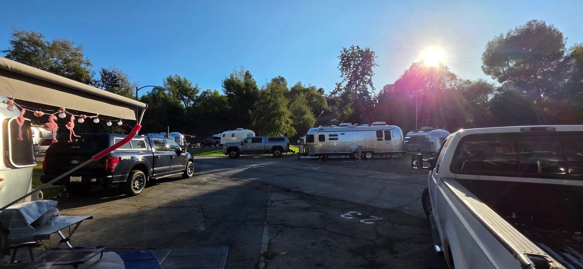 Campground with trailers and trucks, sun in sky. Trees in background, blue sky.