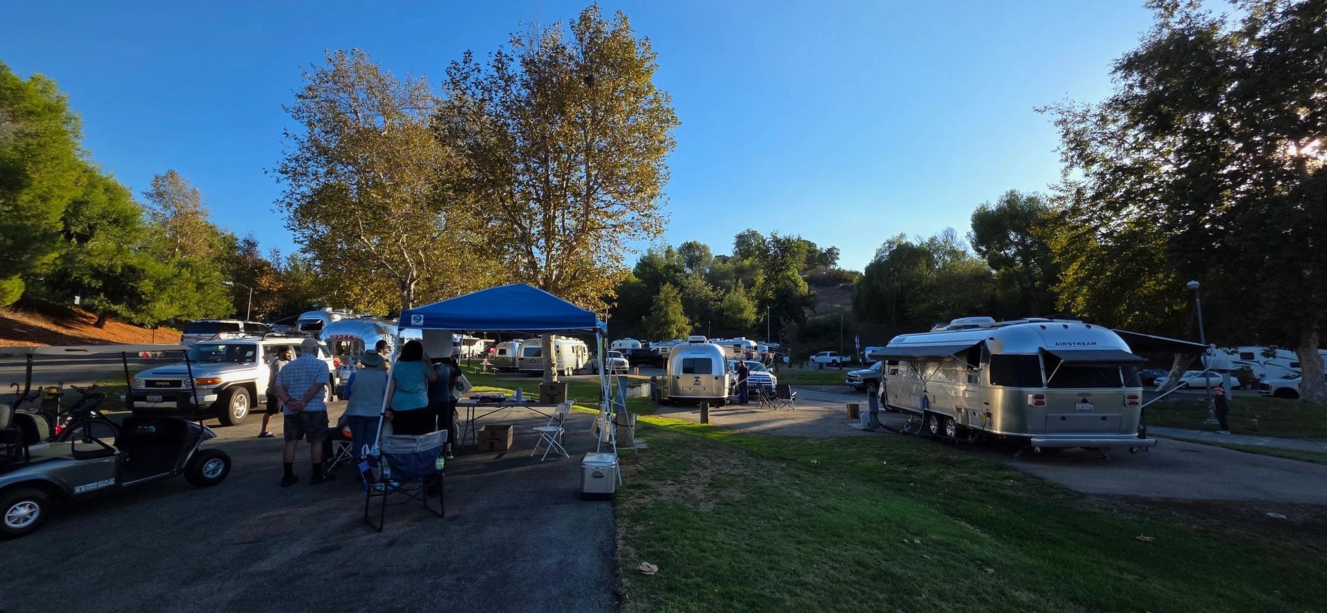 Campground scene with trailers, people, golf carts, and a blue canopy under a clear blue sky.