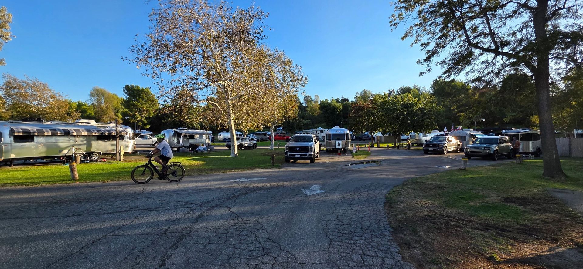 A person rides a bike through a campground with RVs, trees, and a paved road on a sunny day.