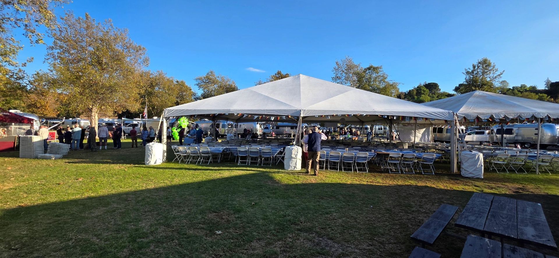 Outdoor event with tents, tables, and people gathered on a grassy field under a blue sky.