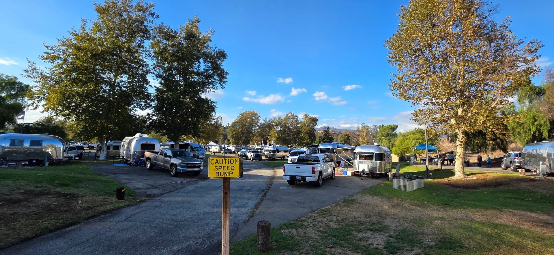 Campground with silver RVs, trees, blue sky, and a road sign.