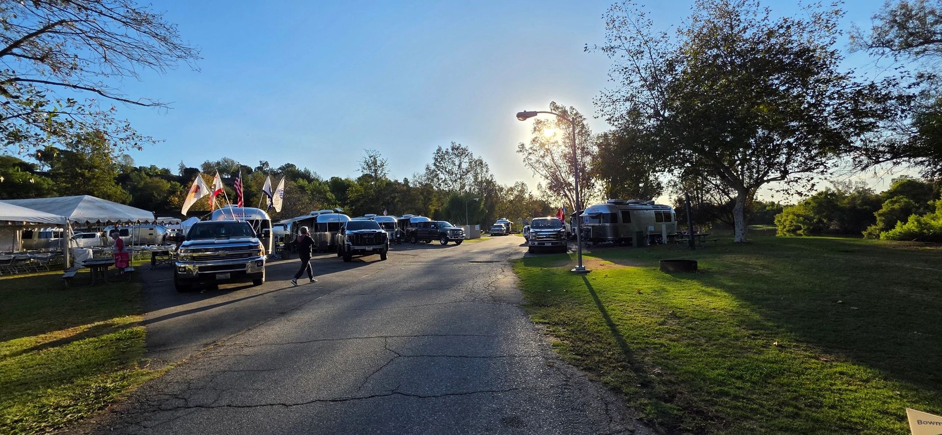 Road lined with parked trucks, trees, and tents on the left, with the sun shining bright.